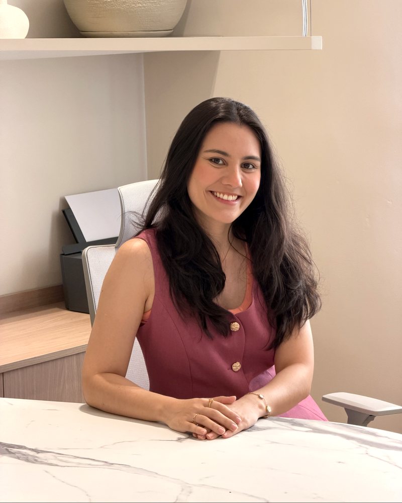 Dra. Roberta Sayuri, médica, de vestido rosa, sorrindo, sentada à mesa do consultório com as mãos cruzadas