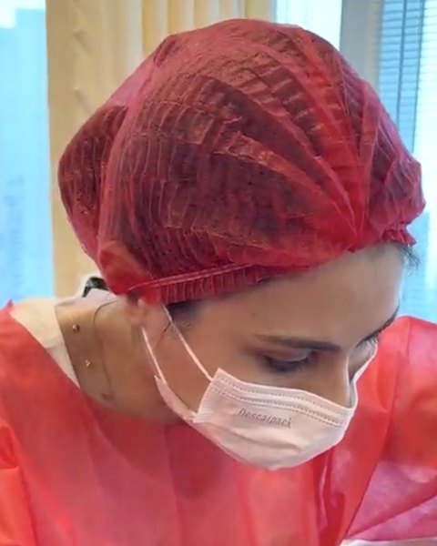 Dra. Roberta Sayuri concentrada durante um procedimento, de avental vermelho, touca e máscara descartáveis
