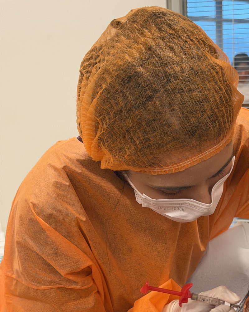 Dra. Roberta Sayuri, de avental, touca e máscara descartáveis, durante um procedimento injetável na face, com luvas